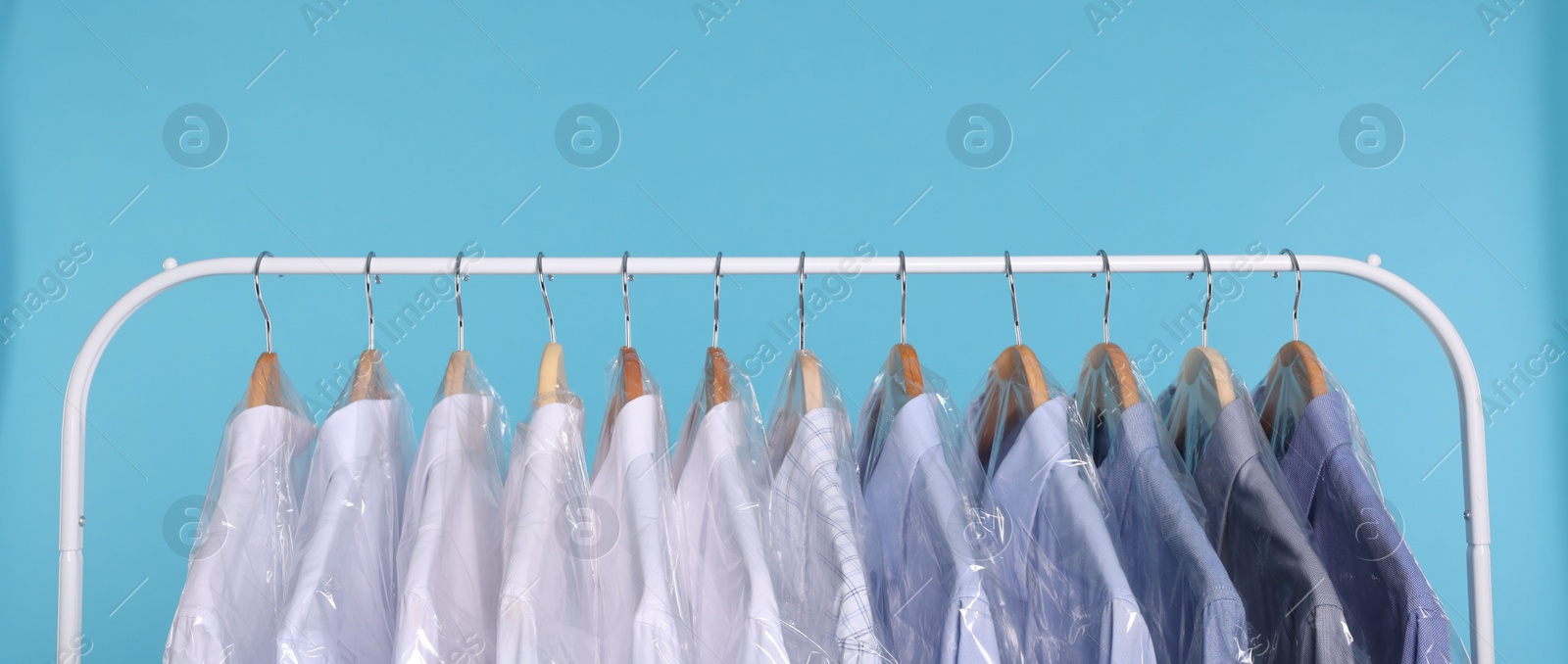 Photo of Dry-cleaning service. Many different clothes in plastic bags hanging on rack against light blue background