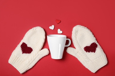 Knitted mittens, cup and paper hearts on red background, flat lay