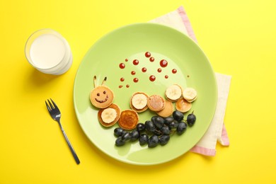 Photo of Creative serving for kids. Plate with cute caterpillar made of pancakes, grapes and banana on yellow background, flat lay