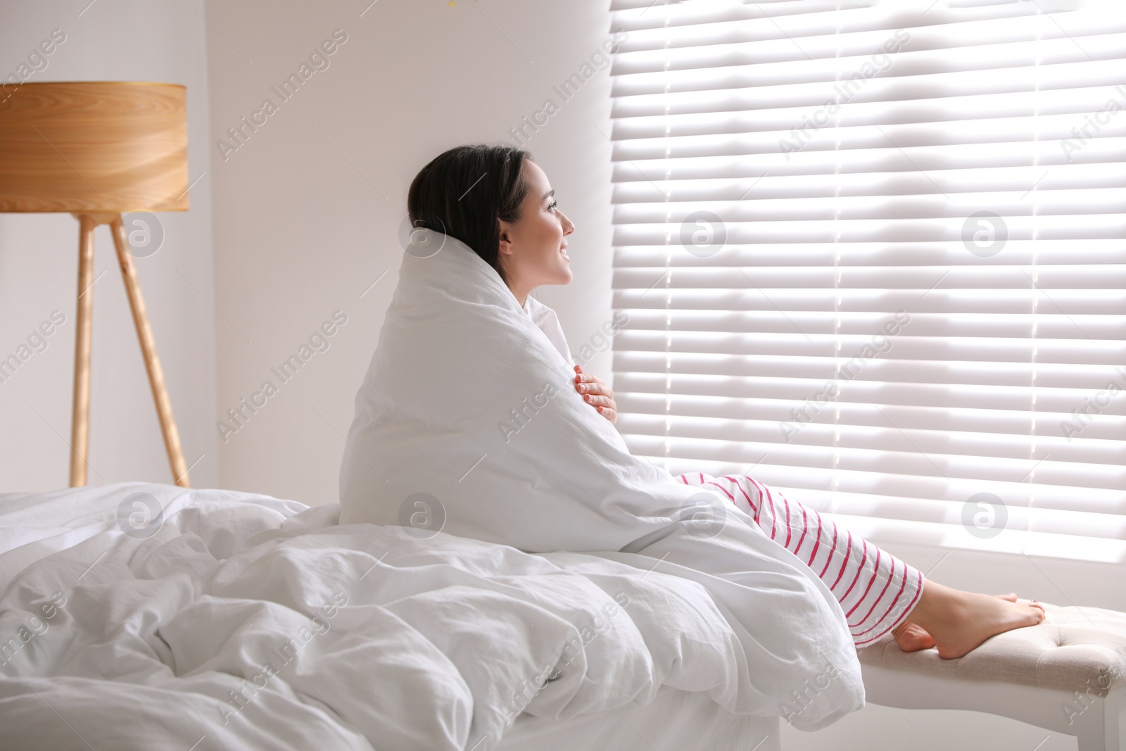 Photo of Beautiful young woman covered with warm blanket near window at home