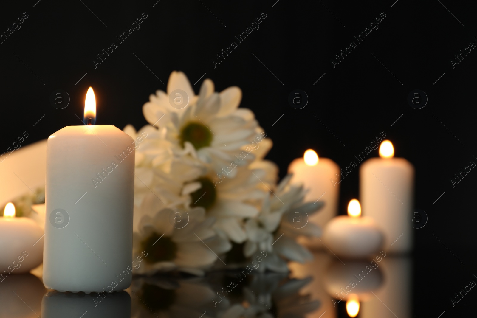 Photo of White chrysanthemum flowers and burning candles on black mirror surface in darkness, space for text. Funeral symbols