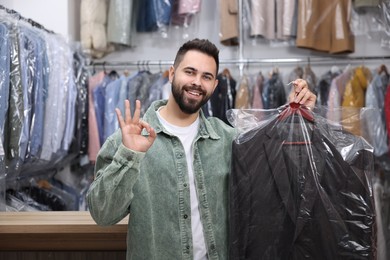 Dry-cleaning service. Happy man holding hanger with jacket in plastic bag and showing ok gesture indoors