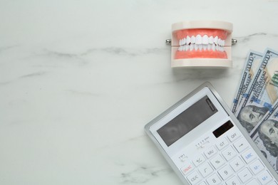 Photo of Educational dental typodont model, dollar banknotes and calculator on white table, flat lay with space for text. Expensive treatment