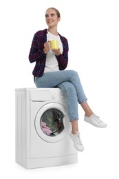 Beautiful young woman with cup of drink on washing machine with laundry against white background