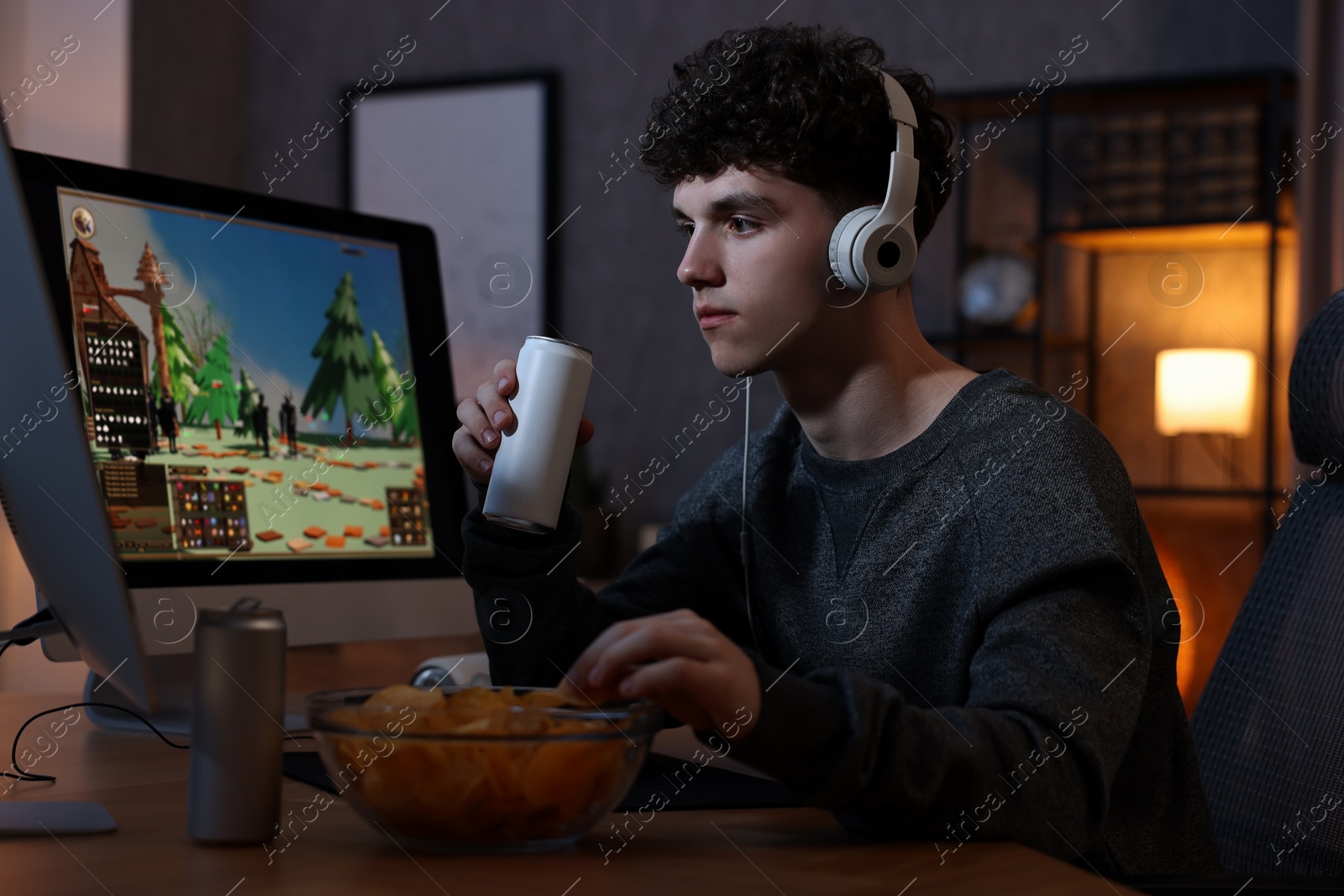 Photo of Young man with energy drink and headphones playing video game at wooden desk indoors