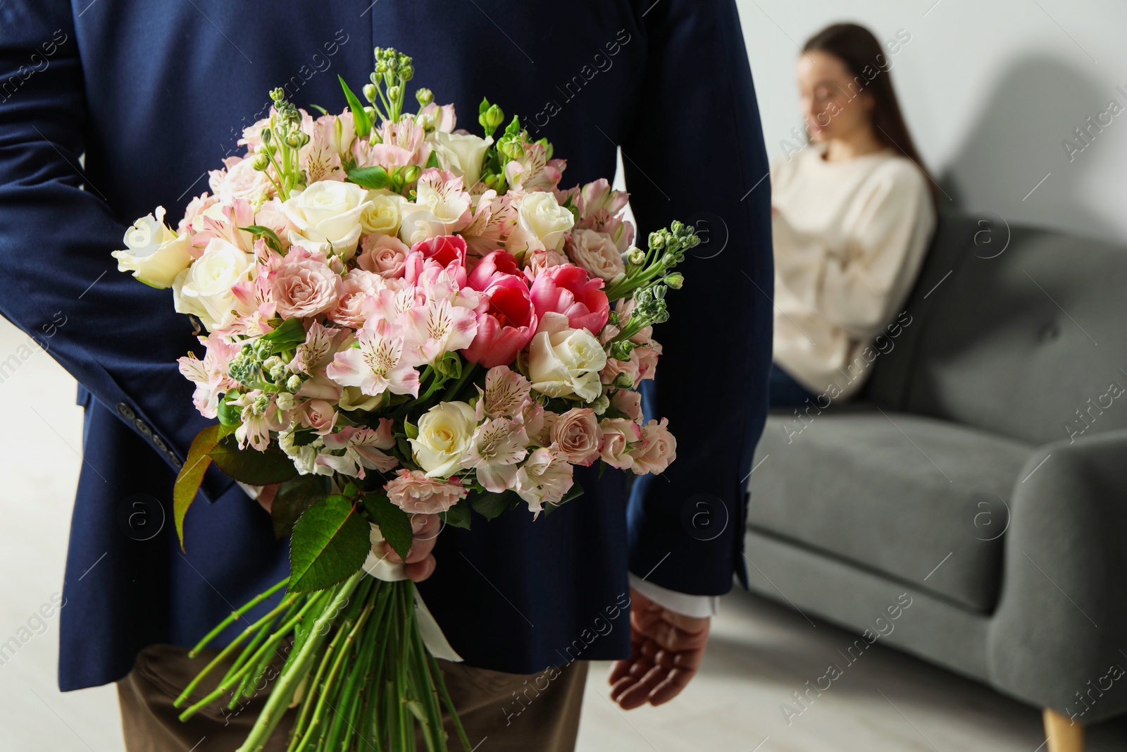 Photo of Man hiding bouquet of flowers for his beloved woman indoors, closeup