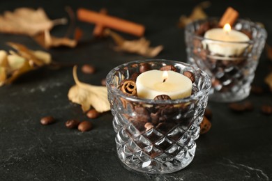 Photo of Glass holders with burning candles, coffee beans and cinnamon on black stone table, closeup