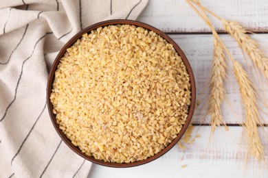 Photo of Raw bulgur in bowl and spikes on white wooden table, flat lay