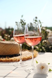 Glasses of rose wine, straw hat and beautiful flower on white wooden table outdoors