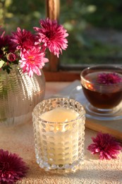 Photo of Burning scented candle in glass holder, cup of tea and chrysanthemum flowers on beige textured table. Autumn season