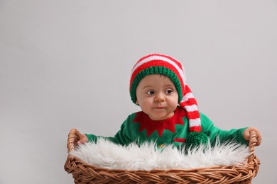 Cute baby in elf costume near wicker basket on light grey background, space for text. Christmas celebration
