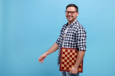 Smiling man holding chessboard on light blue background, space for text