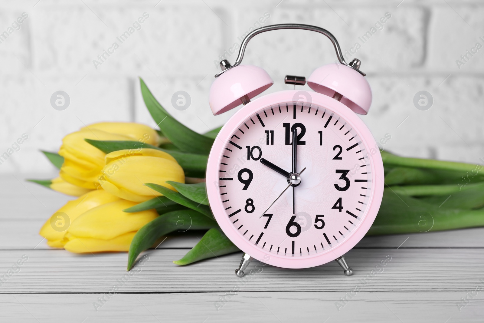 Photo of Pink alarm clock and beautiful tulips on white wooden table against brick wall, closeup. Spring time