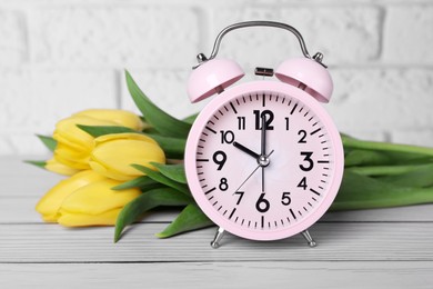 Photo of Pink alarm clock and beautiful tulips on white wooden table against brick wall, closeup. Spring time