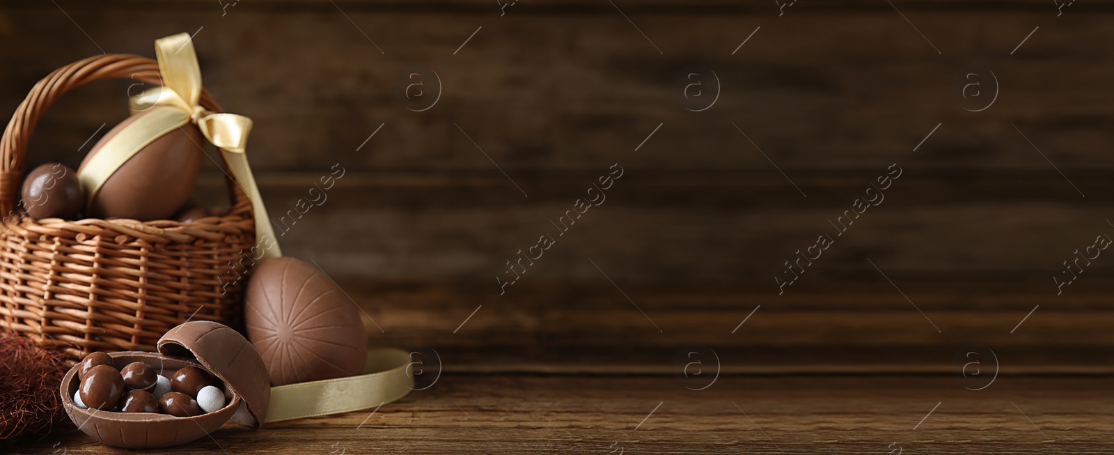 Image of Tasty chocolate eggs and wicker basket on wooden table, space for text. Banner design