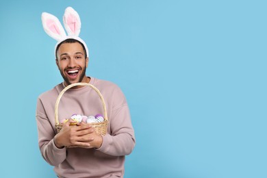Happy African American man in bunny ears headband holding wicker basket with Easter eggs on light blue background, space for text