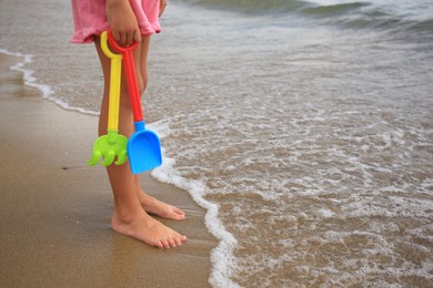 Photo of Little girl with plastic toys standing on sandy beach near sea, closeup. Space for text