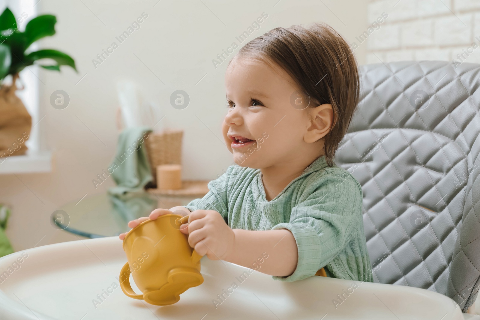 Photo of Cute little baby with cup in high chair indoors