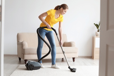 Photo of Young woman cleaning carpet with vacuum in living room