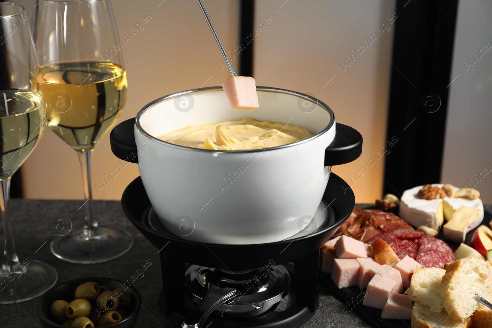 Photo of Dipping piece of ham into fondue pot with melted cheese at grey table with wine and snacks, closeup