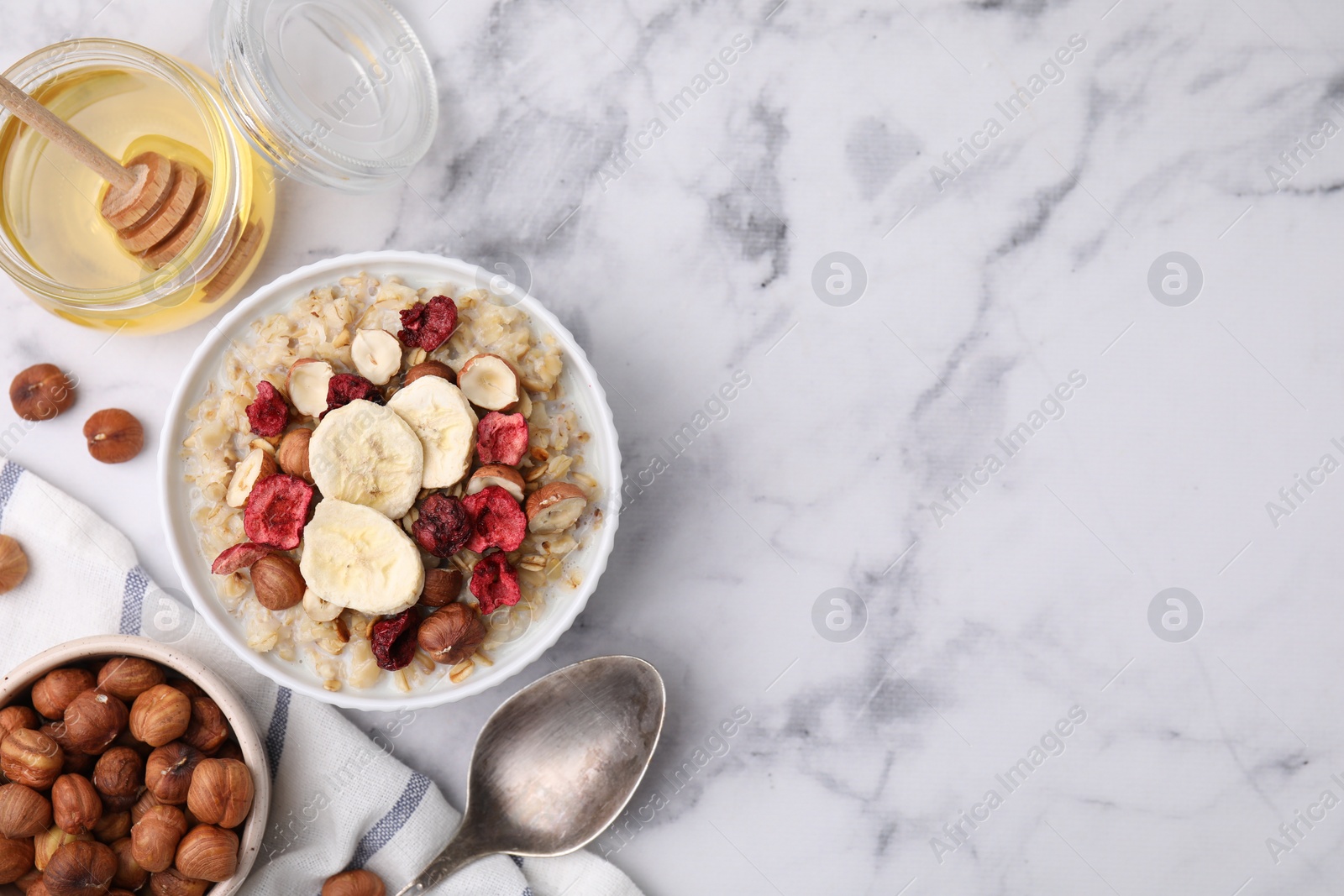 Photo of Delicious oatmeal with freeze dried berries, banana and hazelnuts served on white marble table, flat lay. Space for text