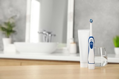 Electric toothbrush, tube with paste and glass of water on wooden table in bathroom. Space for text