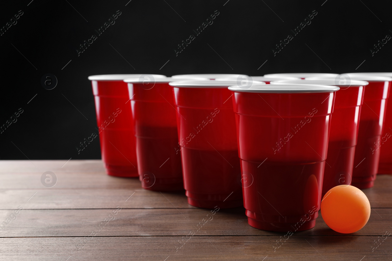 Photo of Plastic cups and ball for beer pong on wooden table against black background
