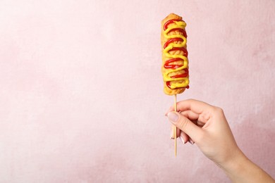 Photo of Woman holding delicious corn dog with mustard and ketchup on pink background, closeup. Space for text