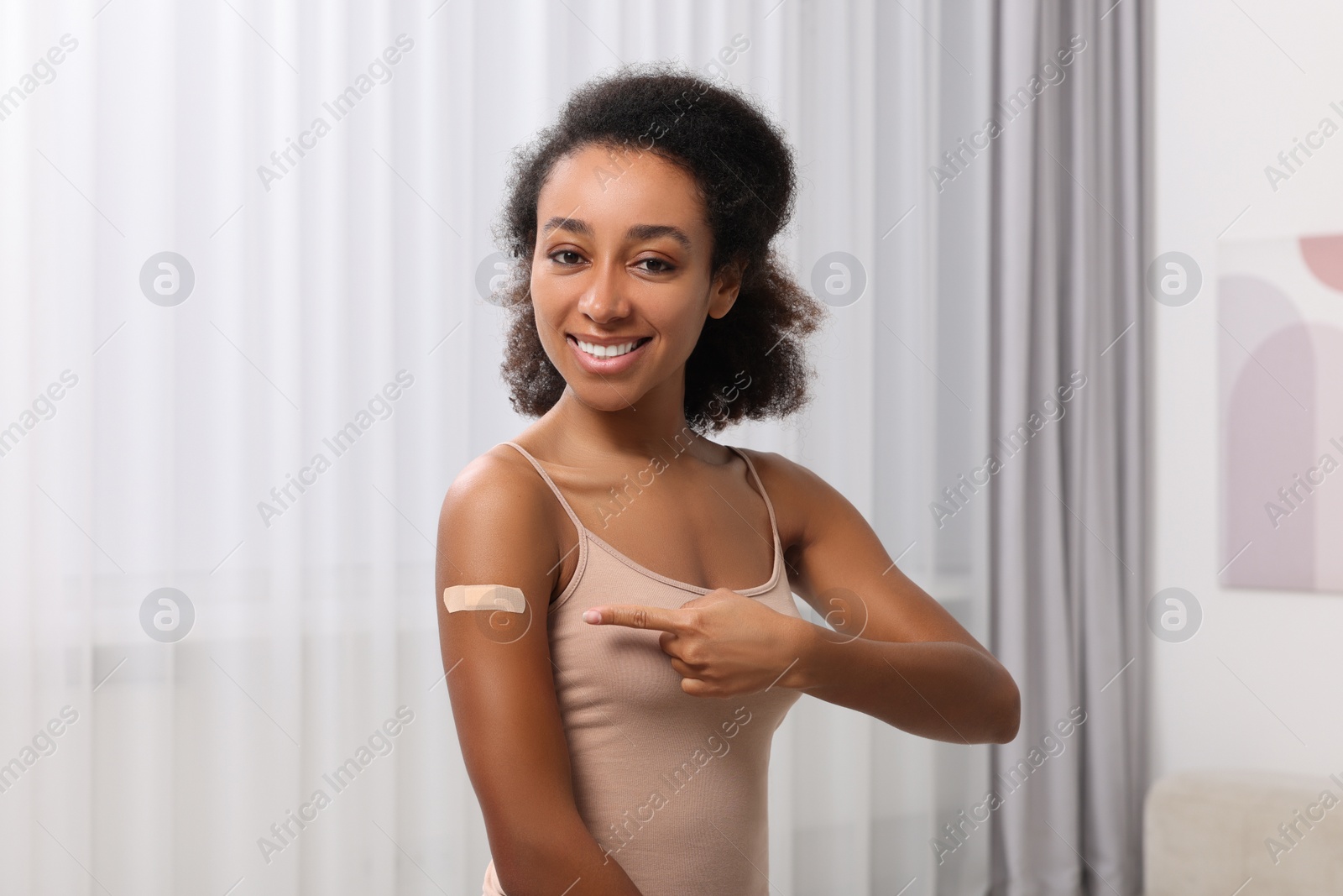 Photo of Happy young woman pointing at adhesive bandage after vaccination indoors