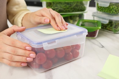 Photo of Woman sticking paper note onto container with fresh cherry tomatoes at white marble table, closeup. Food storage