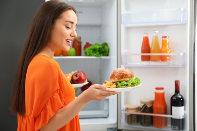 Woman choosing between fruits and burger with French fries near refrigerator in kitchen