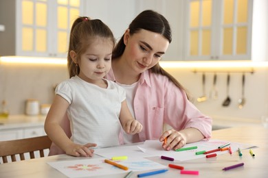 Mother and her little daughter drawing with colorful markers at table in kitchen