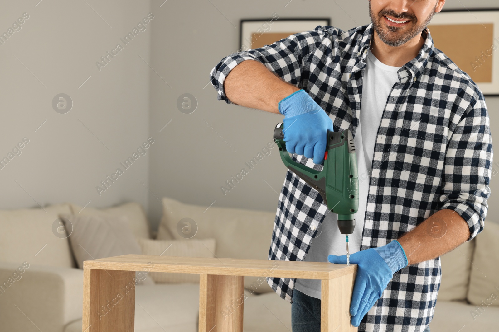 Photo of Man with electric screwdriver assembling wooden furniture at home, closeup. Space for text