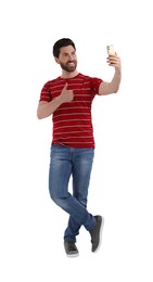 Smiling man taking selfie with smartphone and showing thumbs up on white background