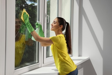 Photo of Happy young woman cleaning window glass with sponge cloth and spray indoors