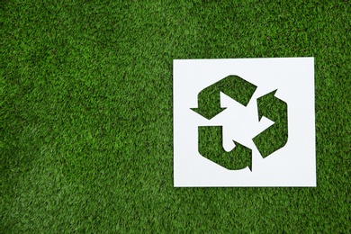 Photo of Sheet of paper with cutout recycling symbol on green grass, top view. Space for text