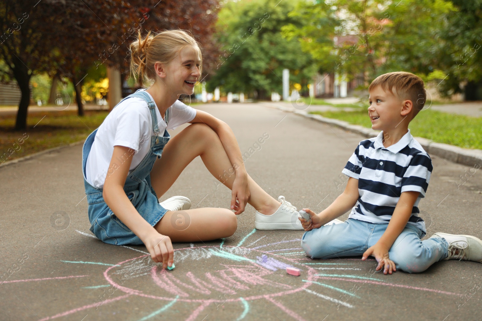 Photo of Teen nanny and cute little boy drawing sun with chalks on asphalt