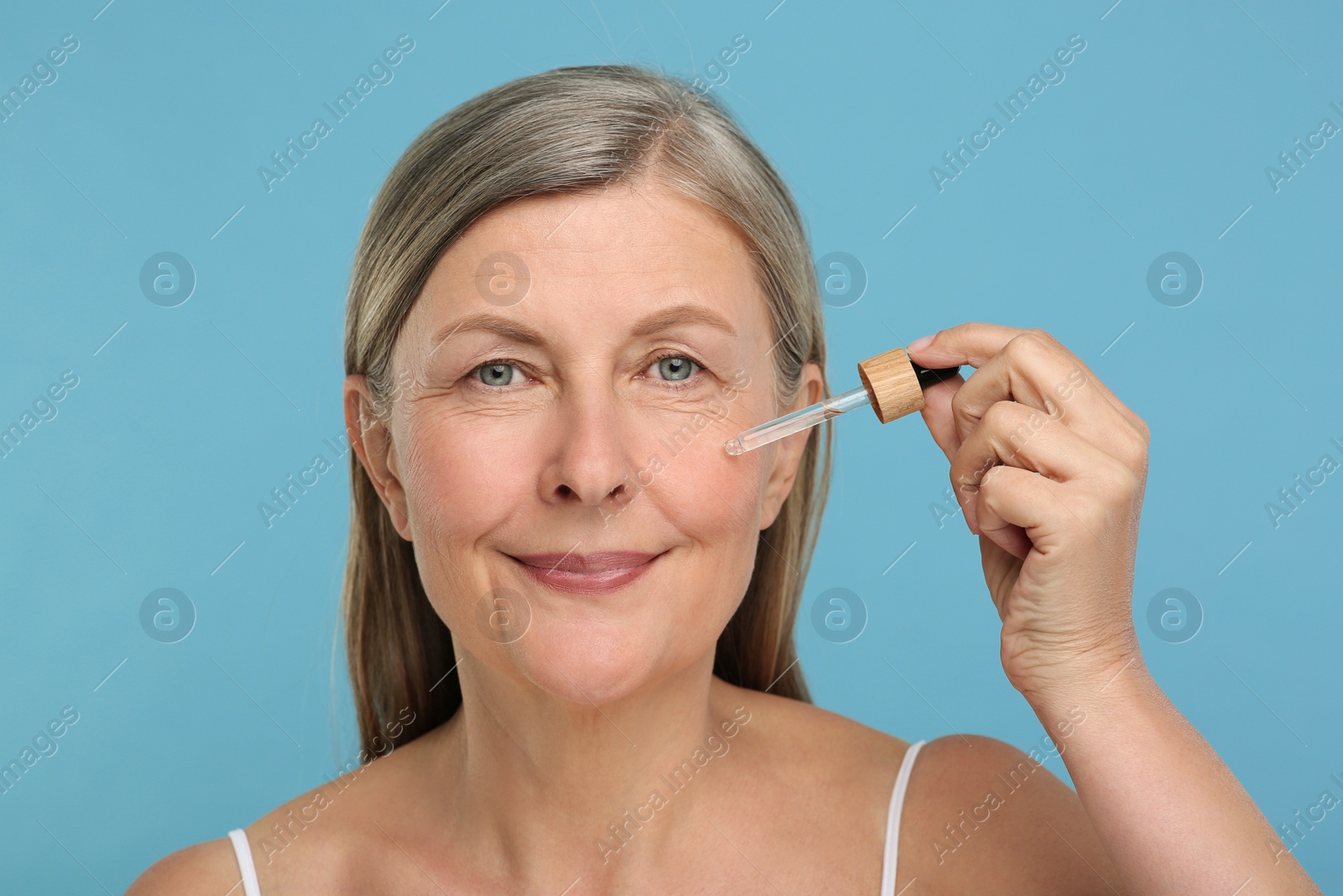 Photo of Senior woman applying cosmetic product on her aging skin against light blue background. Rejuvenation treatment