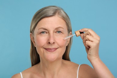 Photo of Senior woman applying cosmetic product on her aging skin against light blue background. Rejuvenation treatment