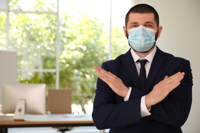 Photo of Man in protective face mask showing stop gesture in office. Prevent spreading of coronavirus