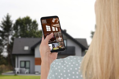 Image of Woman using smart home security system on mobile phone near house outdoors. Device showing different rooms through cameras