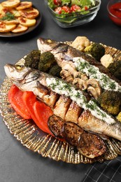 Plate with delicious baked sea bass fish and sauce on black table, closeup