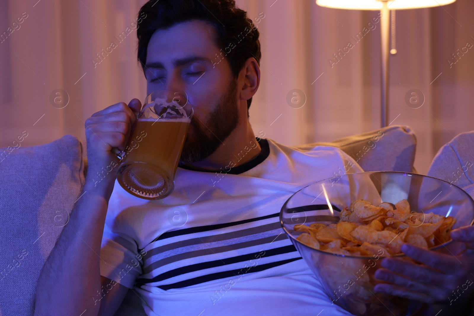 Photo of Man with chips and glass of beer on sofa at night. Bad habit
