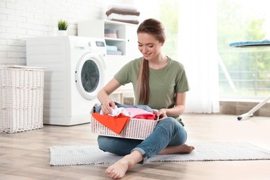 Photo of Young woman with basket of clean laundry on floor in room