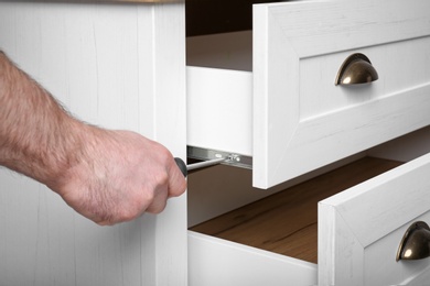 Photo of Man fixing drawer of white wardrobe with screwdriver, closeup