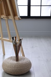 Photo of Different brushes on wicker pouf near easel in artist's studio, space for text