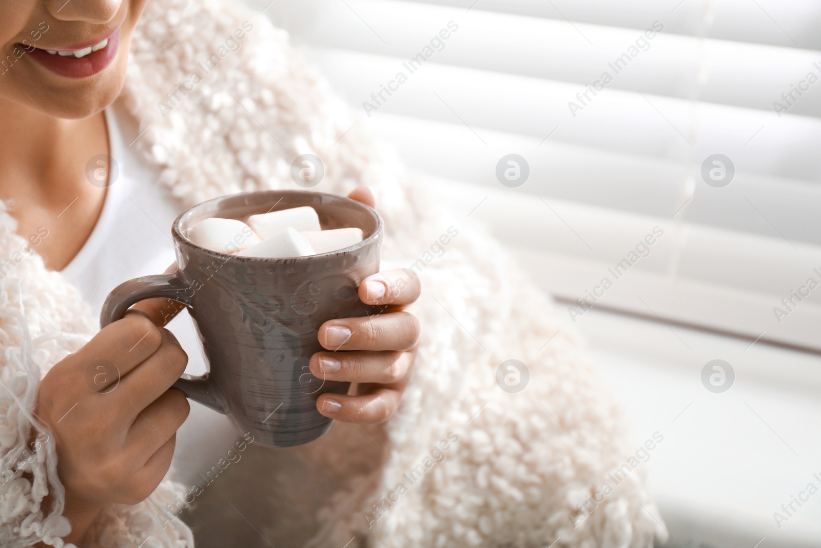 Photo of Young woman with cup of hot drink and marshmallow near window at home, closeup. Winter atmosphere
