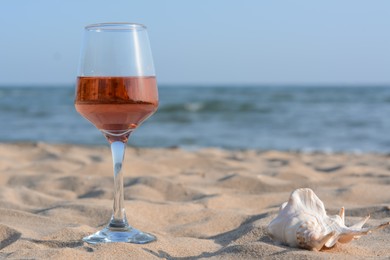 Photo of Glass of tasty rose wine and seashell on sand near sea, space for text