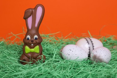 Easter celebration. Funny chocolate bunny and painted eggs with feathers on grass against red background