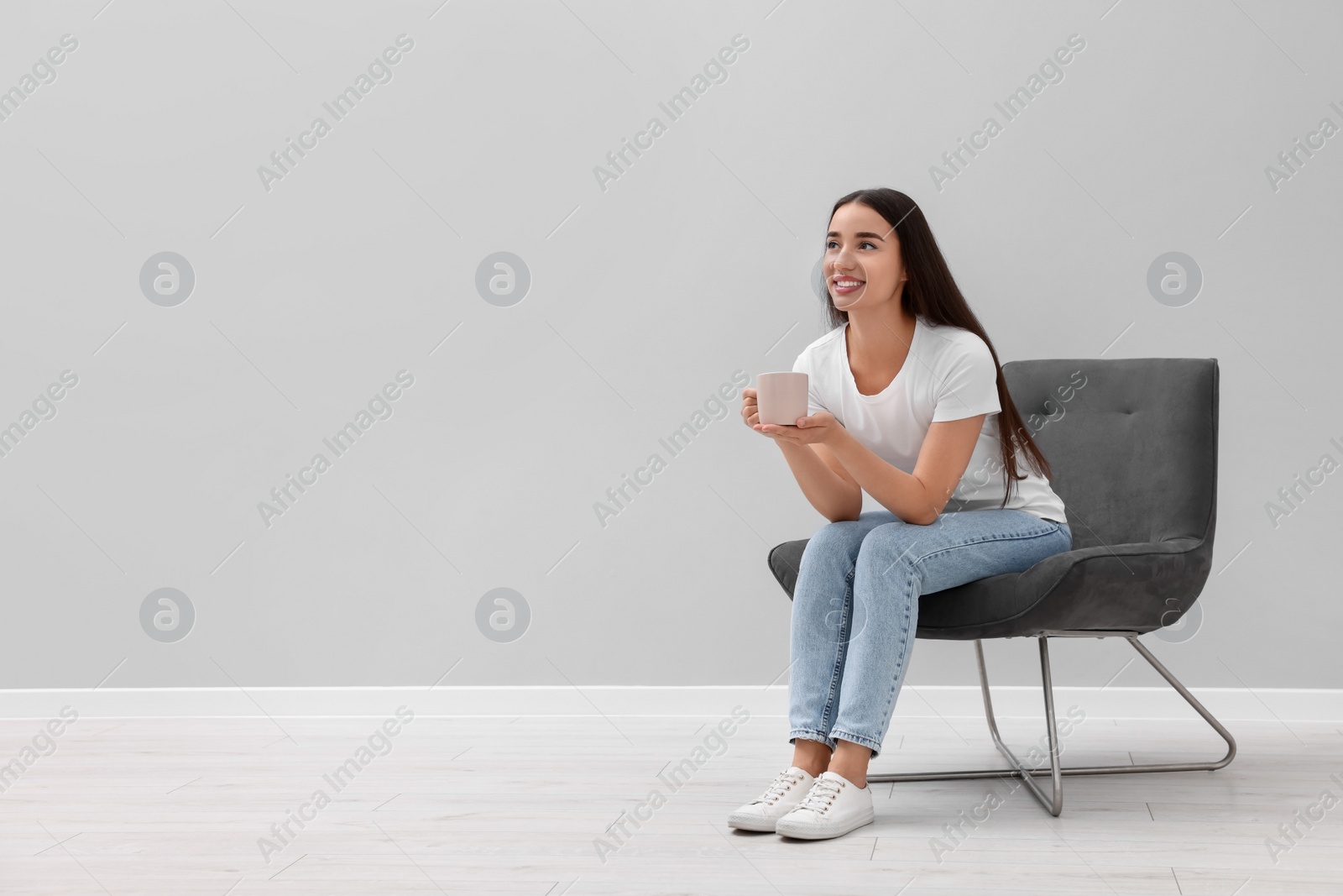 Photo of Beautiful woman with cup of drink sitting in armchair near light grey wall indoors, space for text